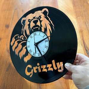 May include: Black vinyl record clock featuring a grizzly bear design in orange. The clock face is white with black Roman numerals and hands. The word "Grizzly" is written in orange script below the clock face.