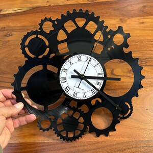 May include: Black gear-shaped wall clock with a white clock face featuring black Roman numerals and hands. The clock is made from a recycled vinyl record and has a unique industrial design. The clock is approximately 30 cm in diameter.