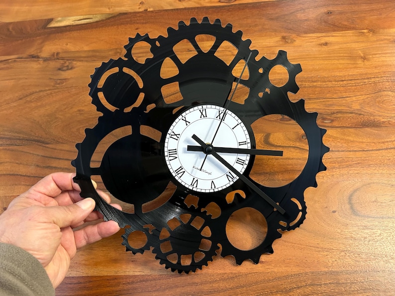 May include: Black gear-shaped wall clock with a white clock face featuring black Roman numerals and hands. The clock is made from a recycled vinyl record and has a unique industrial design. The clock is approximately 30 cm in diameter.