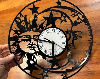 Puede incluir: Reloj de vinilo negro con un diseño de sol y luna. La esfera del reloj es blanca con números romanos negros y agujas negras. El reloj tiene un diámetro de 30,5 cm.