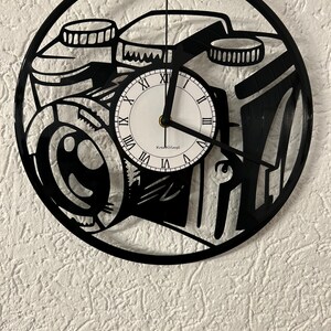 Puede incluir: Reloj de vinilo negro con diseño de cámara. La esfera del reloj es blanca con números romanos negros y el texto "KreativVinyl".
