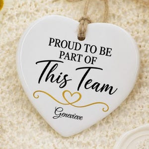 Op de afbeelding: Een wit hartvormig keramisch ornament met de zwarte tekst "PROUD TO BE PART OF THIS TEAM" en de naam "Genevieve" in cursief schrift, voorzien van een gouden hartje. Het hangt aan een bruin touwtje en is omringd door witte en gele bloemen.