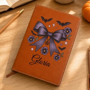 May include: A tan notebook featuring a large purple bow illustration with bats, small flowers, stars, and a pentagram charm. The name Gloria is printed in black script below the bow. The notebook has stitched edges and a ribbon bookmark.
