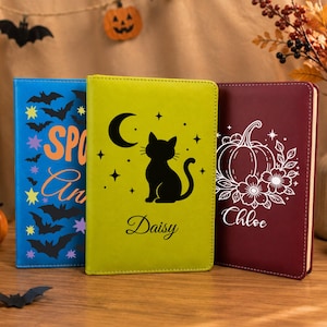 May include: Three rectangular notebooks with distinct covers: a blue one with bats and the text Spooky, a lime green one featuring a black cat, moon, stars, and the name Daisy, and a maroon one with a white pumpkin, floral design, and the name Chloe.