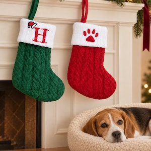 May include: Two cable-knit Christmas stockings hanging from a fireplace mantel. The left stocking is green with a white cuff featuring a red letter H and a small Santa hat. The right stocking is red with a white cuff featuring a red paw print.