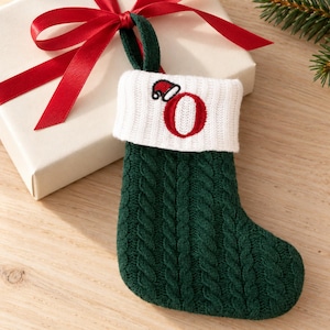 May include: A dark green cable-knit Christmas stocking featuring a white ribbed cuff. The cuff is embroidered with a red letter O topped with a small red and white Santa hat. The stocking includes a matching green loop for hanging purposes.
