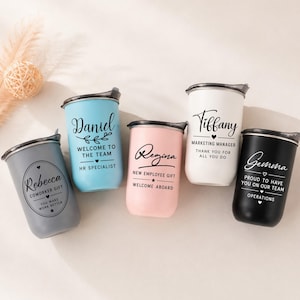 Può includere: Cinque bicchieri in acciaio inossidabile con coperchio in grigio, azzurro, rosa, bianco e nero. Ogni bicchiere presenta nomi e testi stampati personalizzati, tra cui Rebecca, Daniel, Regina, Tiffany e Gemma, con vari titoli professionali.