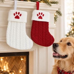 May include: Two cable-knit Christmas stockings, one white and one red, each featuring a white cuff embroidered with a red paw print. The stockings are hung by loops from a mantelpiece, with a golden-colored dog visible in the foreground.