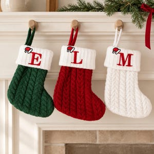 May include: Three cable-knit Christmas stockings in green, red, and white. Each stocking features a white cuff with a red embroidered letter E, L, or M, and a small embroidered Santa hat above the letter. Each has a matching fabric hanging loop.