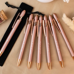 Op de afbeelding: Een verzameling roségouden pennen met gouden accenten, elk gegraveerd met een naam. De pennen worden gepresenteerd met een zwart fluwelen pennenzakje, een kleine glazen buis met gedroogde rozenblaadjes en een vaas met witte madeliefjes.