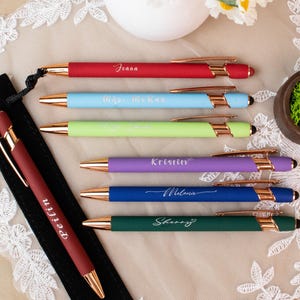 May include: A collection of colorful pens with gold accents, each inscribed with a name. The pens are red, light blue, green, purple, blue, and dark green. A black pen case with a pen is on the left. A small bowl with moss and rings is on the right.