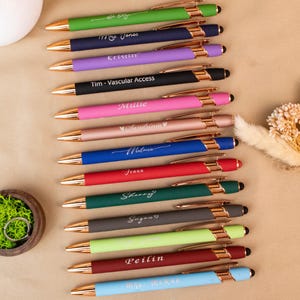 May include: A collection of personalized pens in various colors, including green, navy, purple, black, pink, rose gold, blue, red, dark green, gray, light green, and burgundy. Each pen has a gold-colored accent and is engraved with a name or text.