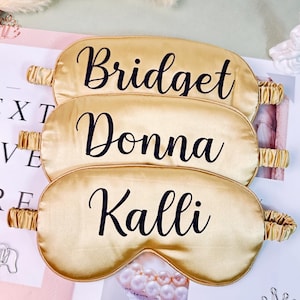 Puede incluir: Tres antifaces de satén dorado con letras negras que dicen "Bridget", "Donna" y "Kalli". Cada antifaz tiene una banda elástica. Los antifaces se muestran en una página de revista con elementos decorativos.