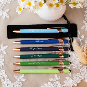 May include: A collection of personalized pens in various colors, including light blue, navy, dark green, and light green. Each pen has a gold-colored tip and is engraved with a name or title. The pens are displayed on a lace surface.