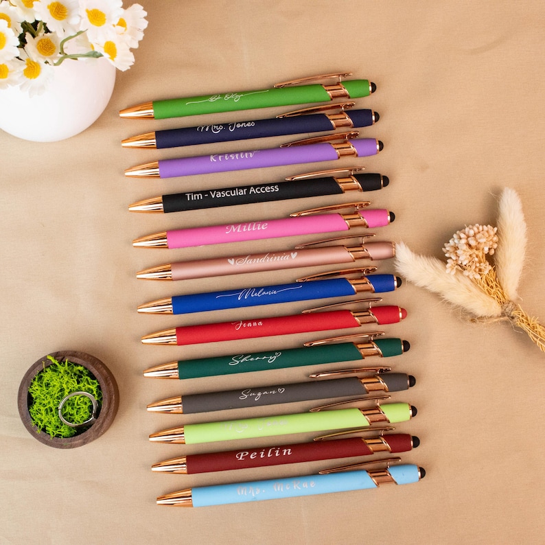 May include: A collection of personalized pens in various colors, including green, navy, purple, black, pink, rose gold, blue, red, dark green, gray, light green, and burgundy. Each pen has a gold-colored accent and is engraved with a name or text.