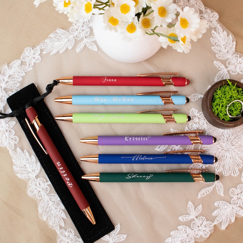 May include: A collection of colorful pens with gold accents, each inscribed with a name. The pens are red, light blue, green, purple, blue, and dark green. A black pen case with a pen is on the left. A small bowl with moss and rings is on the right.