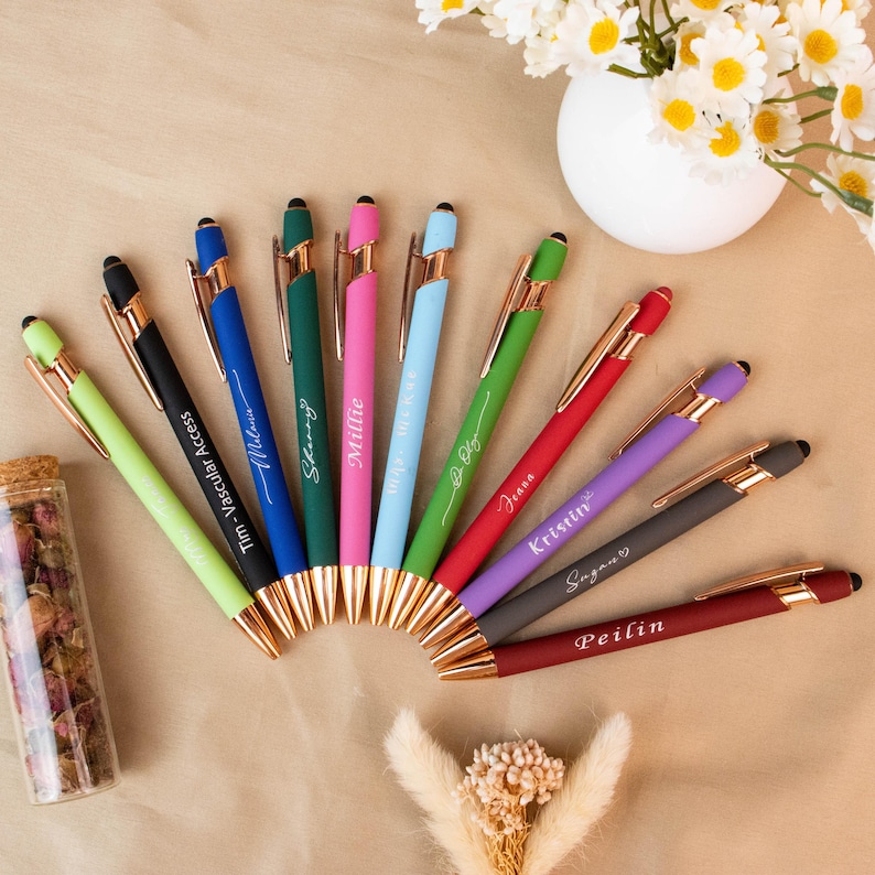 May include: A collection of colorful pens with gold accents, each featuring a different name in a cursive font. The pens are arranged in a fan shape, with a small glass bottle of dried flowers and a vase of daisies in the background.