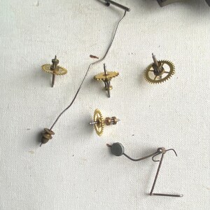 Puede incluir: Primer plano de piezas de reloj desmontadas, incluyendo peque&ntilde;os engranajes de lat&oacute;n, un alambre met&aacute;lico delgado y un peque&ntilde;o p&eacute;ndulo. Los componentes est&aacute;n dispersos sobre una superficie blanca, sugiriendo reparaci&oacute;n o restauraci&oacute;n.
