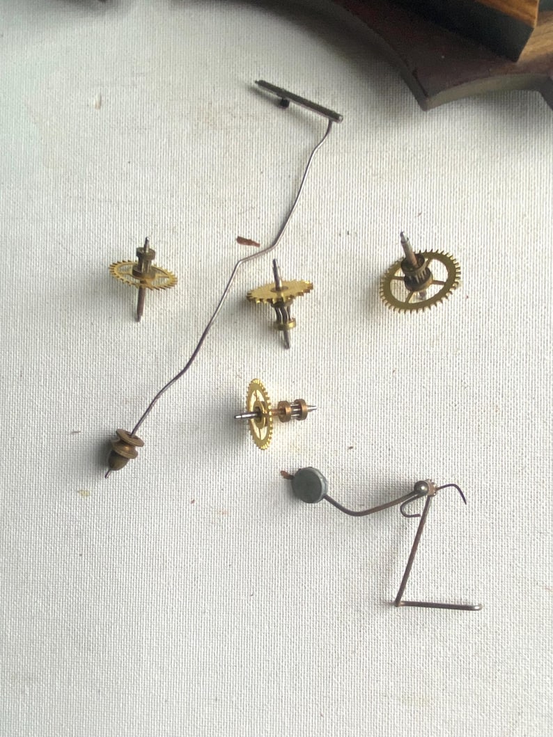 Puede incluir: Primer plano de piezas de reloj desmontadas, incluyendo peque&ntilde;os engranajes de lat&oacute;n, un alambre met&aacute;lico delgado y un peque&ntilde;o p&eacute;ndulo. Los componentes est&aacute;n dispersos sobre una superficie blanca, sugiriendo reparaci&oacute;n o restauraci&oacute;n.