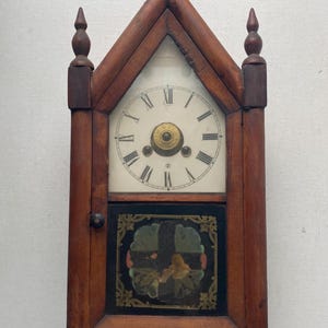 May include: A wooden wall clock with a pointed gable top and two finials. The clock face features black Roman numerals on a white background. Below the face is a rectangular panel with a black background, gold decorative corners, and a central floral motif.