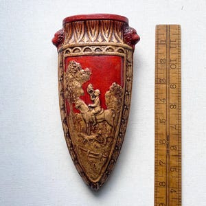 May include: A decorative, shield-shaped wall pocket with a red central panel featuring a relief of a rider on horseback, a dog, and trees. The pocket is approximately 10 inches tall, with a tan and red color scheme and ornate detailing.