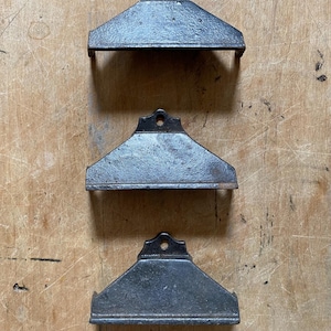 May include: Three antique, cast iron shelf brackets with a dark, weathered finish. Each bracket has a triangular shape with a small hole at the top for mounting. The brackets are displayed on a wooden surface.