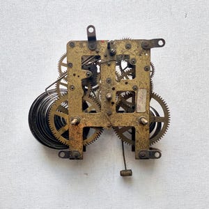 May include: A vintage clock movement with brass gears and a spring mechanism. The intricate gears are arranged within a rectangular frame, with a pendulum attachment at the bottom. The clock mechanism is set against a white background.