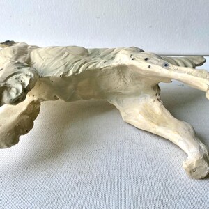 May include: A cream-colored, recumbent dog sculpture. The dog is lying down with its legs extended. The sculpture has a textured surface and is painted with subtle variations in color, giving it a weathered appearance.