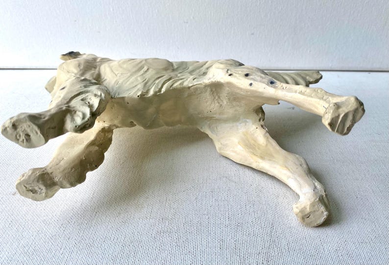 May include: A cream-colored, recumbent dog sculpture. The dog is lying down with its legs extended. The sculpture has a textured surface and is painted with subtle variations in color, giving it a weathered appearance.