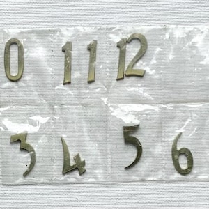 May include: A set of metallic numeral digits zero through twelve, individually encased in a clear plastic grid. The numbers are arranged in two rows, with the top row containing 9, 1, 0, 11, 12 and the bottom row containing 1, 2, 3, 4, 5, 6, 7, 8.