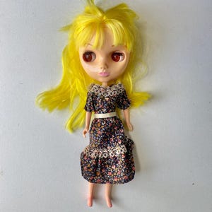 May include: A doll with bright yellow hair and large, dark red eyes. The doll wears a dark blue dress featuring a small floral pattern in orange, white, and blue, accented with white lace trim at the collar and waist, and a white belt at the waist.
