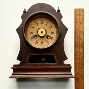 Puede incluir: Reloj de sobremesa de madera antiguo con esfera redonda y n&uacute;meros romanos. El reloj tiene un acabado marr&oacute;n oscuro y un dise&ntilde;o decorativo. Una regla de madera se coloca junto al reloj, mostrando medidas en cent&iacute;metros.