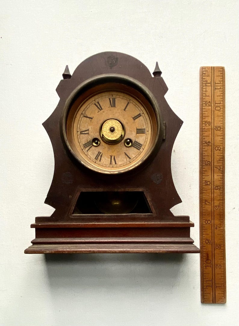 Puede incluir: Reloj de sobremesa de madera antiguo con esfera redonda y n&uacute;meros romanos. El reloj tiene un acabado marr&oacute;n oscuro y un dise&ntilde;o decorativo. Una regla de madera se coloca junto al reloj, mostrando medidas en cent&iacute;metros.
