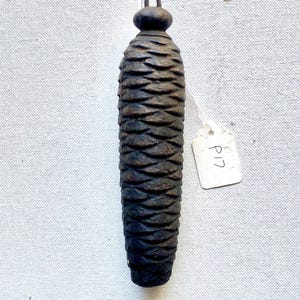 May include: A dark brown, carved wooden pinecone-shaped object with a loop at the top and a small tag attached. The pinecone has a textured, layered design. The tag reads "P17".