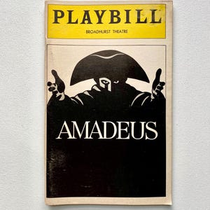 May include: A vintage Playbill from the Broadhurst Theatre for the play "Amadeus." The cover features a black and white illustration of a figure in a tricorn hat, with the title in bold white letters against a black background. The top section is yellow with the word "PLAYBILL."