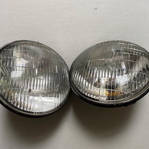 May include: Two vintage, round headlights with a clear, ribbed glass lens and a black metal rim. The headlights are likely for a classic car or motorcycle, offering a retro aesthetic. The lenses are reflective, suggesting they are in good condition.