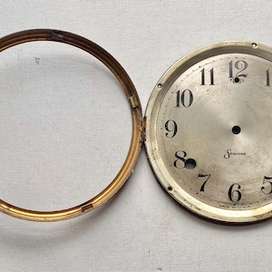 May include: An antique clock face with a gold-colored bezel and a silver-colored face. The face has black numerals and the word "Sessions" printed in the center. The bezel is open, revealing the clock face.