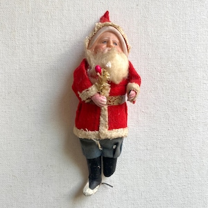May include: Vintage Santa Claus figurine in a red coat with white trim, gray pants, and black boots. The Santa figure has a white beard, a red hat, and is holding a small gold and pink decoration. A classic Christmas decoration.