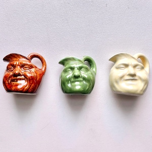 May include: Three small, ceramic pitchers with human face designs. The pitchers are in shades of orange, green, and cream. Each pitcher has a handle and a spout. The faces have smiling expressions.