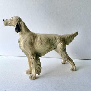 May include: A cream-colored ceramic dog figurine in a standing pose. The dog has black spots and black ear detailing. The figurine has a textured appearance and is set against a white background. The dog's tail is slightly raised.