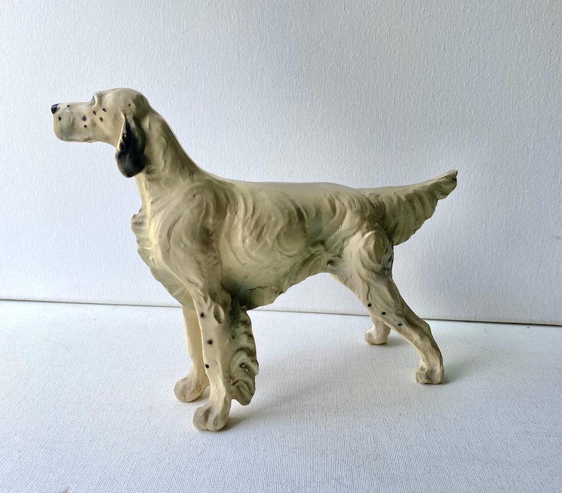 May include: A cream-colored ceramic dog figurine in a standing pose. The dog has black spots and black ear detailing. The figurine has a textured appearance and is set against a white background. The dog's tail is slightly raised.