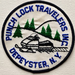 May include: Circular patch with the text "PUNCH LOCK TRAVELERS INC DEPEYSTER N.Y." in blue embroidery. The patch features a black and white snowmobile, green pine trees, and a black mountain range on a white background, with a black border.