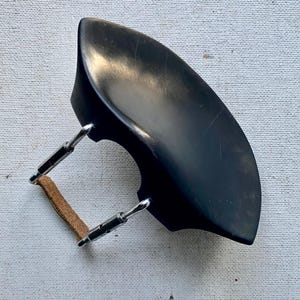 May include: A black violin chin rest featuring a smooth, curved surface and two silver-toned metal mounting brackets. The brackets are connected by a tan leather strap, providing a secure attachment mechanism for the instrument component.