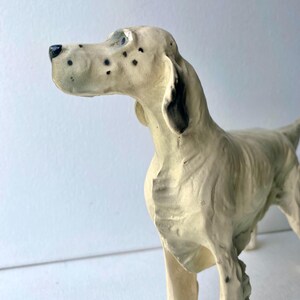 May include: A cream-colored dog figurine with black spots and a black nose. The dog is standing, with its head turned to the left. The figurine has detailed features and a textured surface.