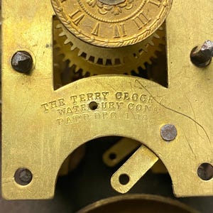 Puede incluir: Primer plano de un mecanismo de reloj antiguo con engranajes y placas de lat&oacute;n. El texto "THE TERRY CLOCK CO. WATERBURY CONN. PATD. DEC. 1 1863" es visible. El intrincado dise&ntilde;o del reloj muestra su edad y artesan&iacute;a.