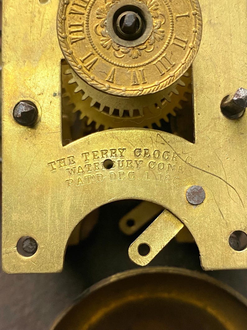 Puede incluir: Primer plano de un mecanismo de reloj antiguo con engranajes y placas de lat&oacute;n. El texto "THE TERRY CLOCK CO. WATERBURY CONN. PATD. DEC. 1 1863" es visible. El intrincado dise&ntilde;o del reloj muestra su edad y artesan&iacute;a.