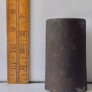 May include: A dark brown, cylindrical object with a rough, textured surface. A wooden ruler is positioned vertically next to the object, showing it stands approximately 4.5 inches tall. The object has a flat top and a flat base.