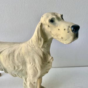 May include: A cream-colored dog figurine with a detailed, textured surface. The dog has a long snout with black spots and a black nose. The figurine is standing, with its head turned slightly to the right. The dog's eyes are dark.