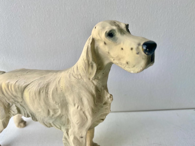 May include: A cream-colored dog figurine with a detailed, textured surface. The dog has a long snout with black spots and a black nose. The figurine is standing, with its head turned slightly to the right. The dog's eyes are dark.