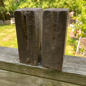 May include: Two rectangular, dark brown, weathered cast iron blocks with a rough, pitted texture. Each block features a vertical groove running down the center of the front face. The objects have a solid, heavy appearance with sharp edges.